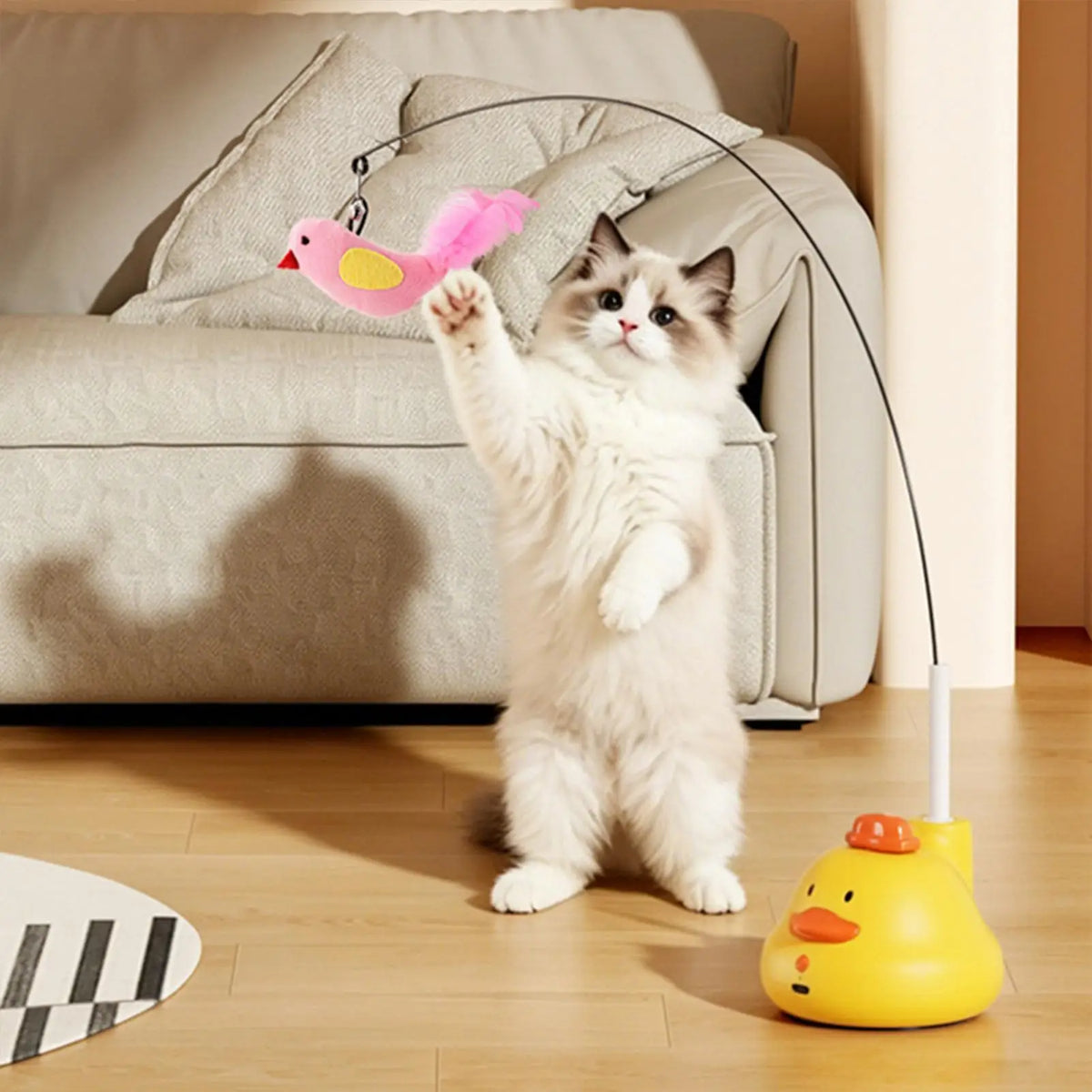 A fluffy white cat plays with a pink feather toy and a yellow duck-shaped interactive feeder.