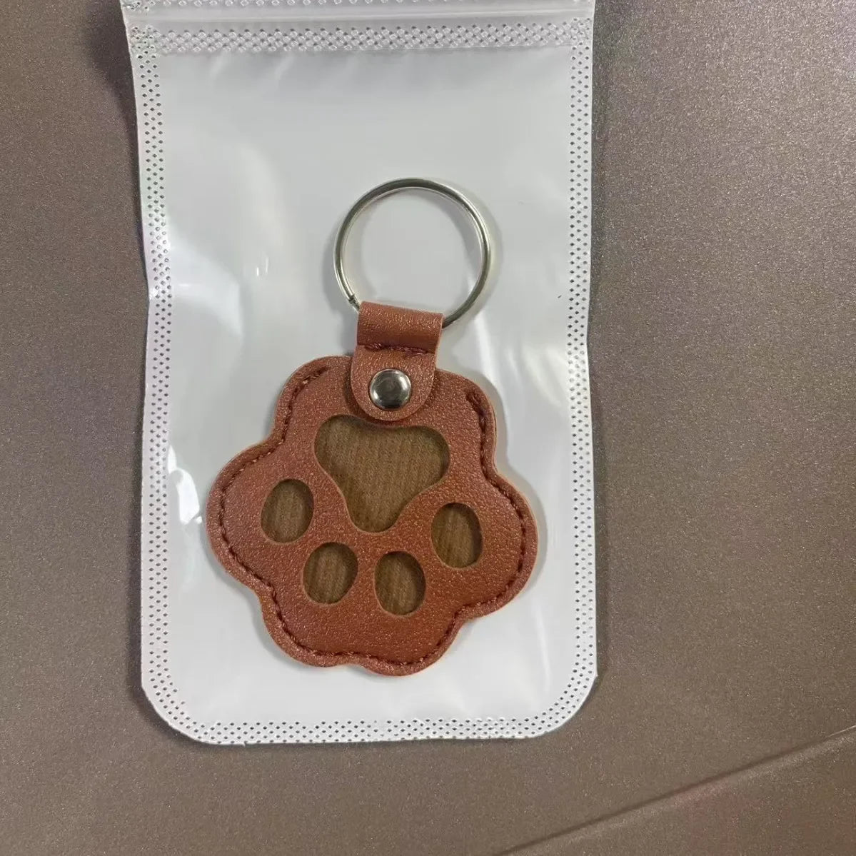 A round, light brown leather pet paw print keychain charm is displayed inside a clear plastic pouch.