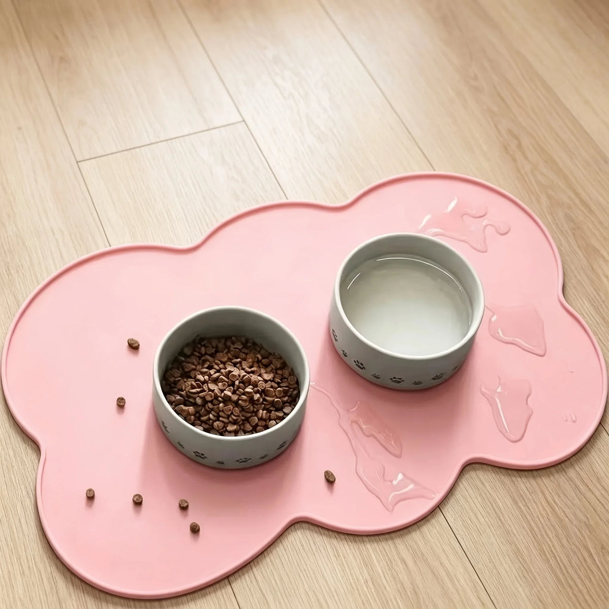 A pink silicone pet feeding mat with a raised edge holds a small bowl of dry food and a white liquid in another bowl.