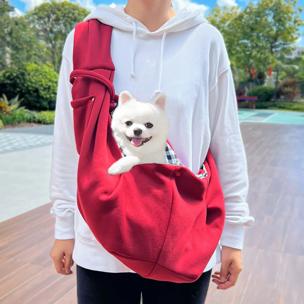 A white dog is carried in a red hands-free pet sling carrier worn by a person.