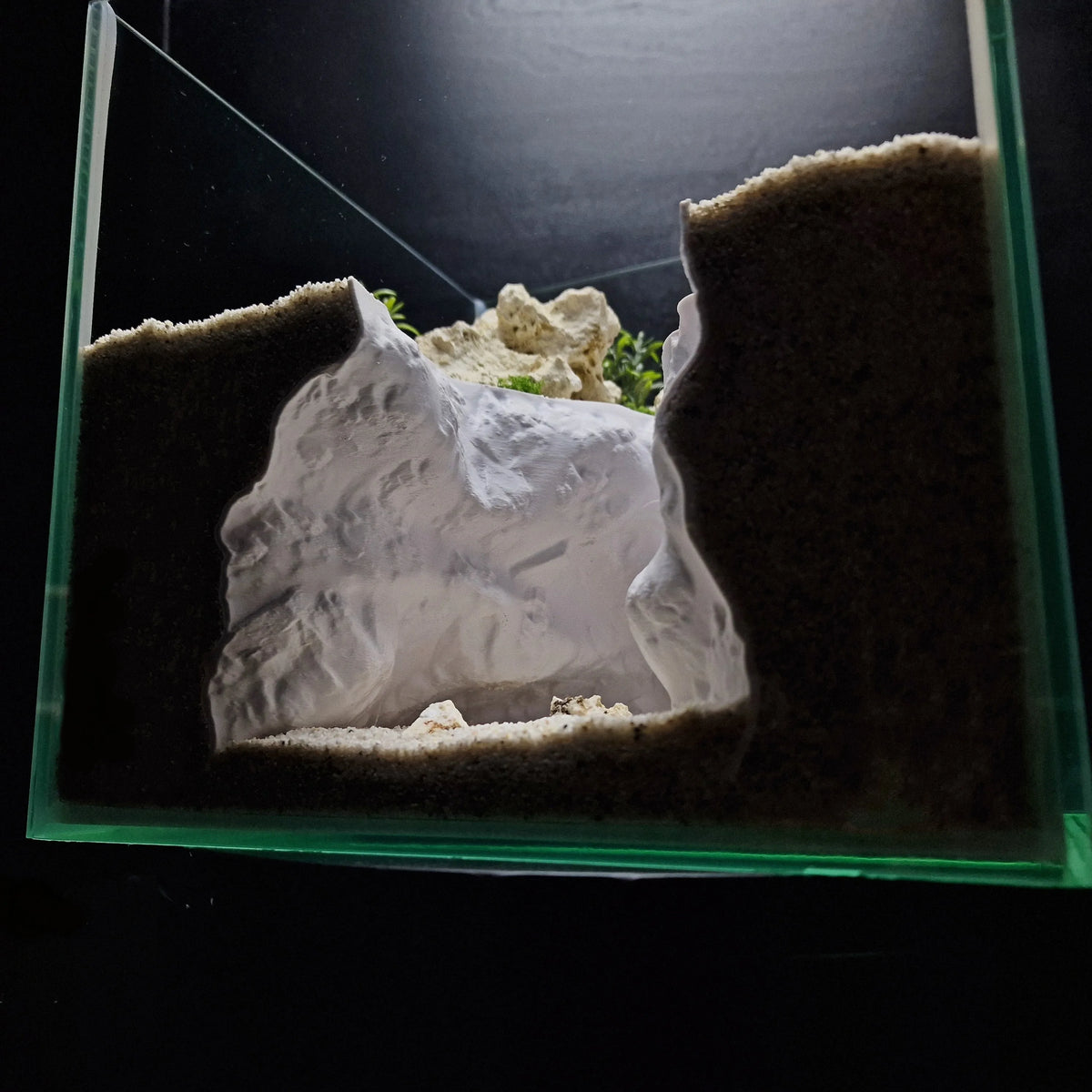 A clear glass aquarium features a 3D rock cave ornament with natural terrain design and sand.