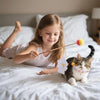 A young girl is holding a stick with a pink pom-pom attached, next to a cat wearing an adorable pink bow collar.