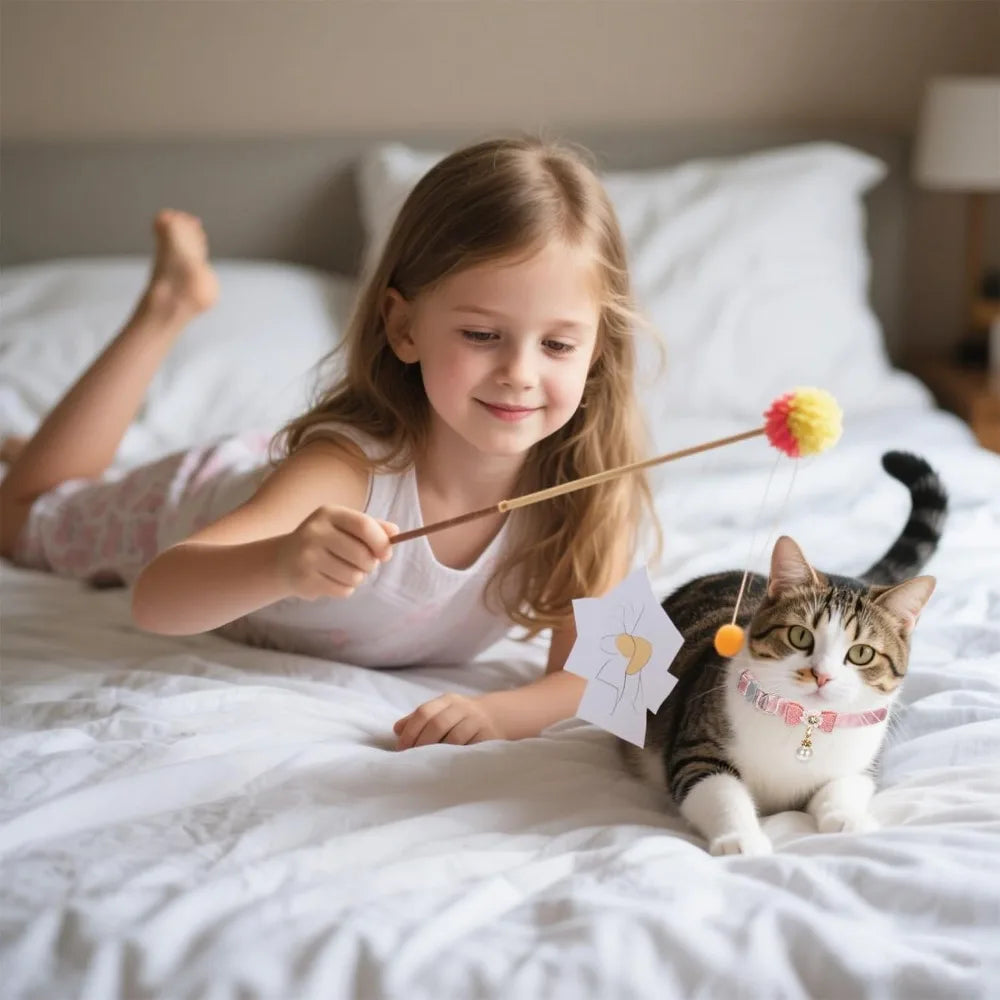 A young girl is holding a stick with a pink pom-pom attached, next to a cat wearing an adorable pink bow collar.