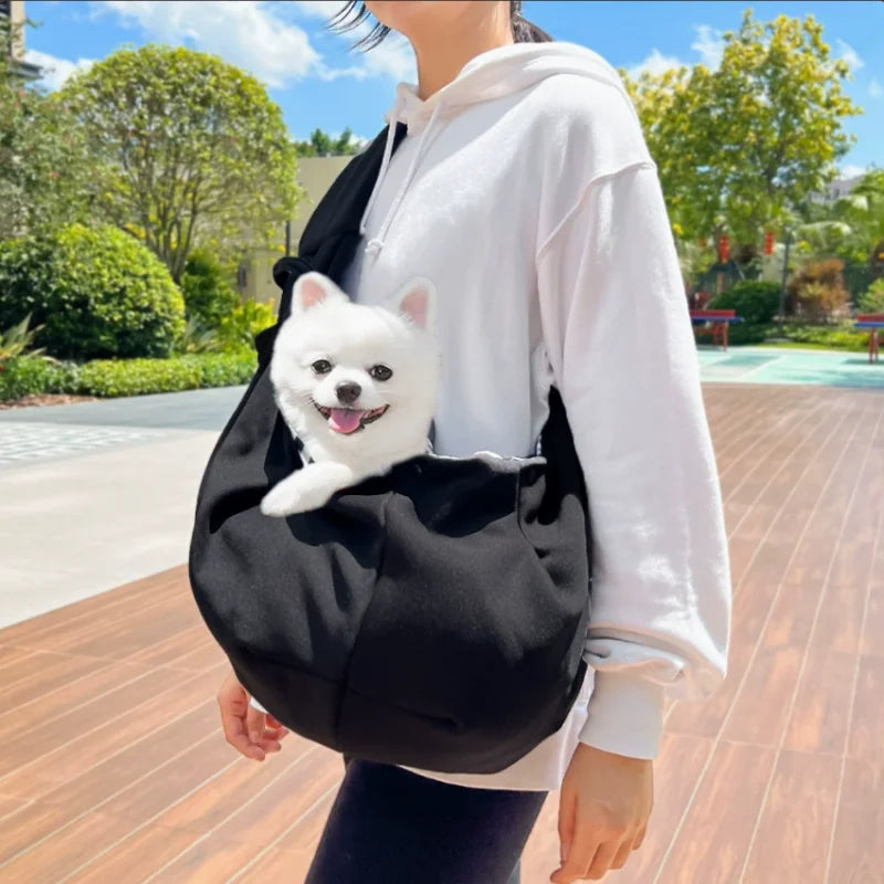 A person wearing a white hoodie is carrying a black pet sling carrier with a small white dog in it.