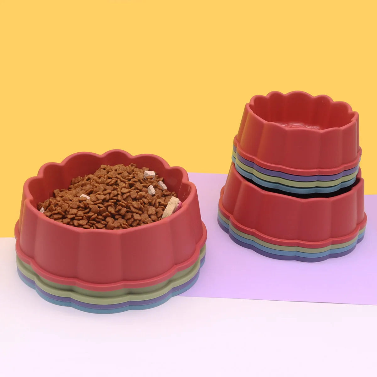 A stack of three colorful, non-slip pet feeding bowls in shades of red and green on a light purple surface.