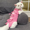 A white cat wearing a pink striped post-operative recovery vest lies on a gray sofa.