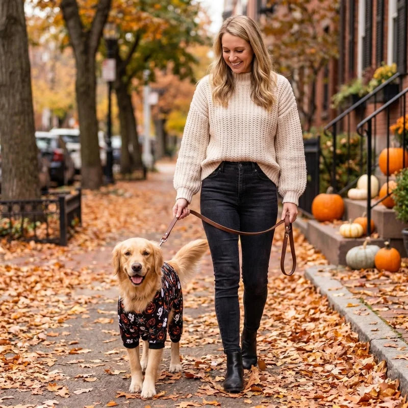 A woman is walking a golden retriever through a path covered in autumn leaves wearing a cream knit sweater and dark jeans.