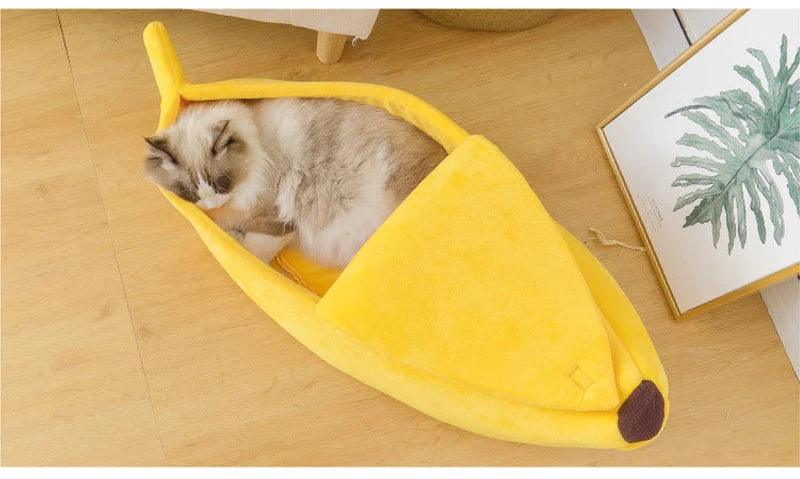 Un petit chat tigré dort dans un lit pour animaux en forme de banane jaune sur un plancher en bois.