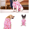 A dog wearing a pink patterned recovery suit sits on a white rug.