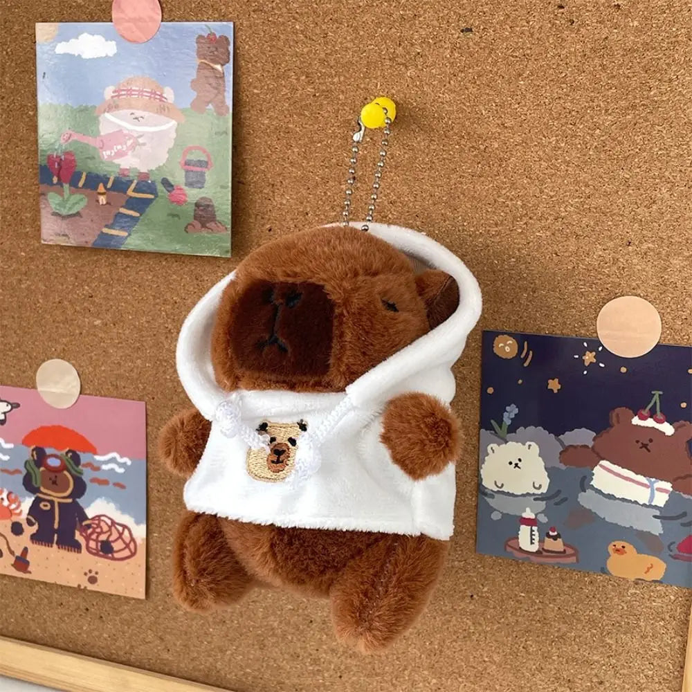 Braune Capybara-Plüschpuppe mit weißem Hoodie und goldenem Anhänger, auf einer Korkwand
