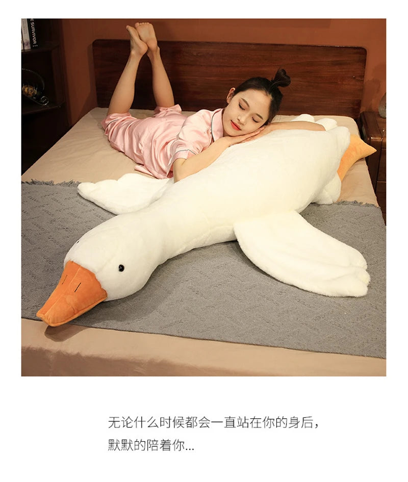 A woman is lying on a bed with a large white plush goose body pillow, looking down.