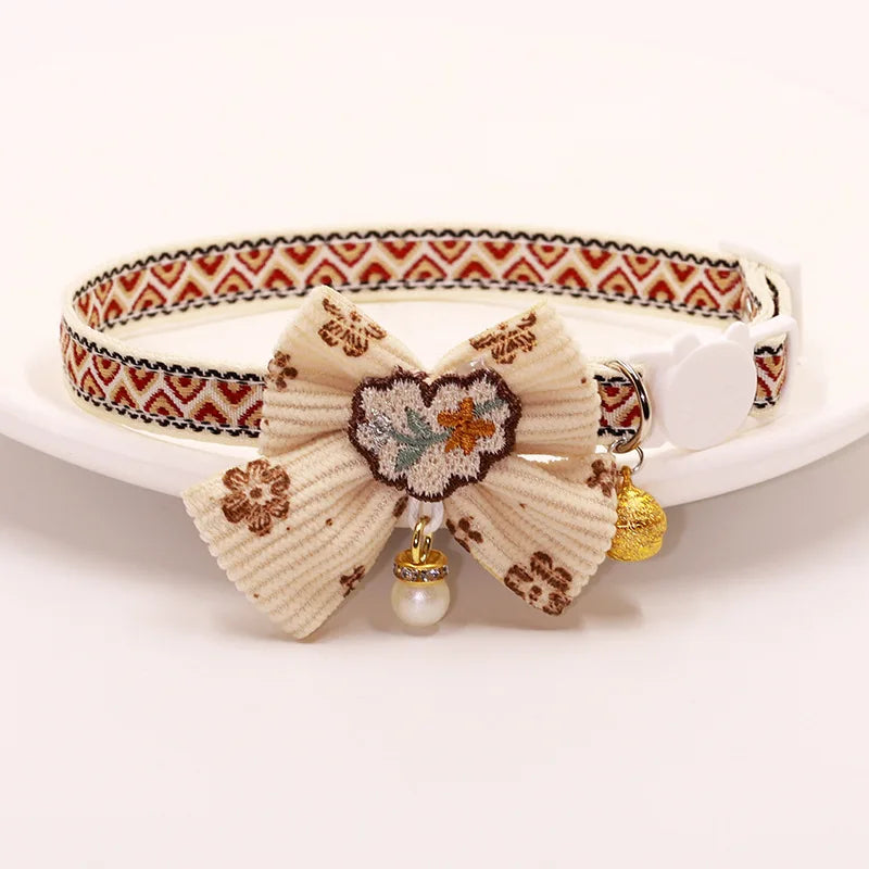 A beige breakaway cat collar featuring a fabric bowknot and a gold bell attached.