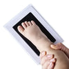 Un kit d'empreinte de main et de pied montrant le pied d'un bébé pressé dans un cadre blanc par la main d'un adulte.