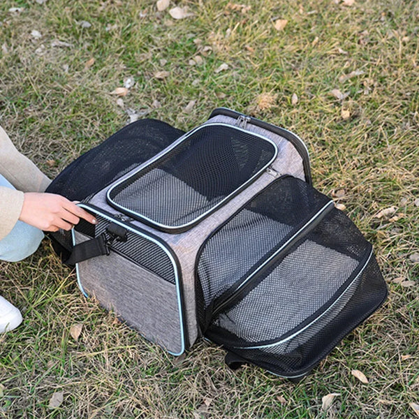 A gray expandable pet carrier backpack with black mesh openings and reflective trim lying on grass.
