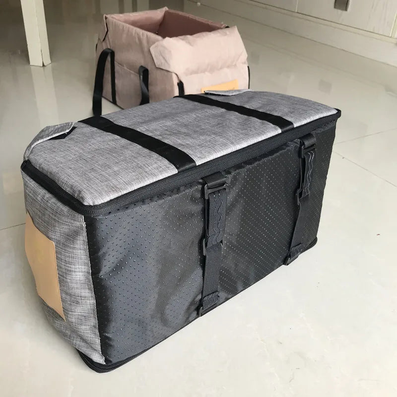 A gray portable pet car seat carrier with black straps and a light pink fabric bag is shown on a floor.