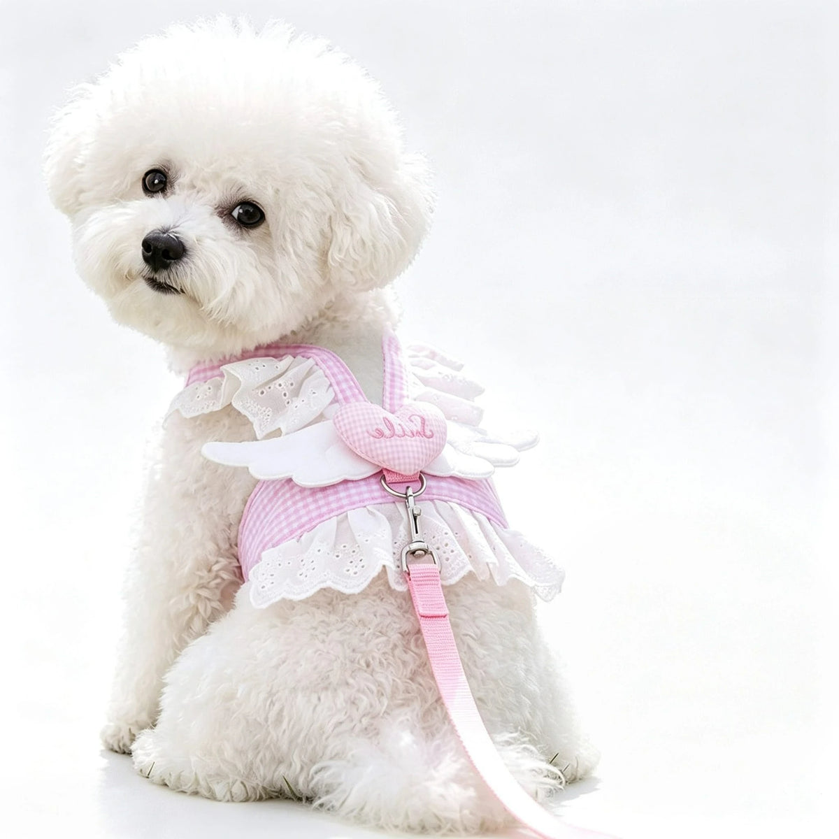 A fluffy white dog wearing a pink lace ruffle plaid harness vest with an angel heart design.