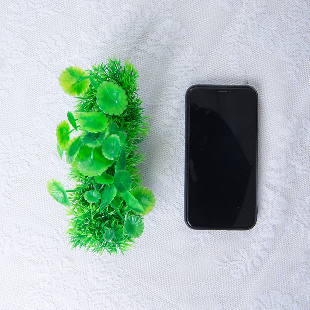 Planta de acuario artificial con hojas y tallos verdes, junto a un smartphone negro sobre una superficie blanca.