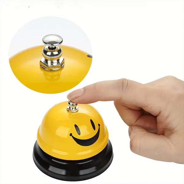 A hand is holding a yellow classic brass call bell with a smiling face and a shiny top.