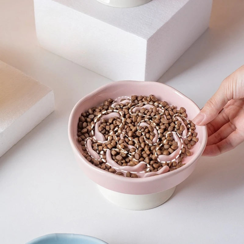 A hand is filling a pink ceramic slow feeder pet bowl with brown kibble.