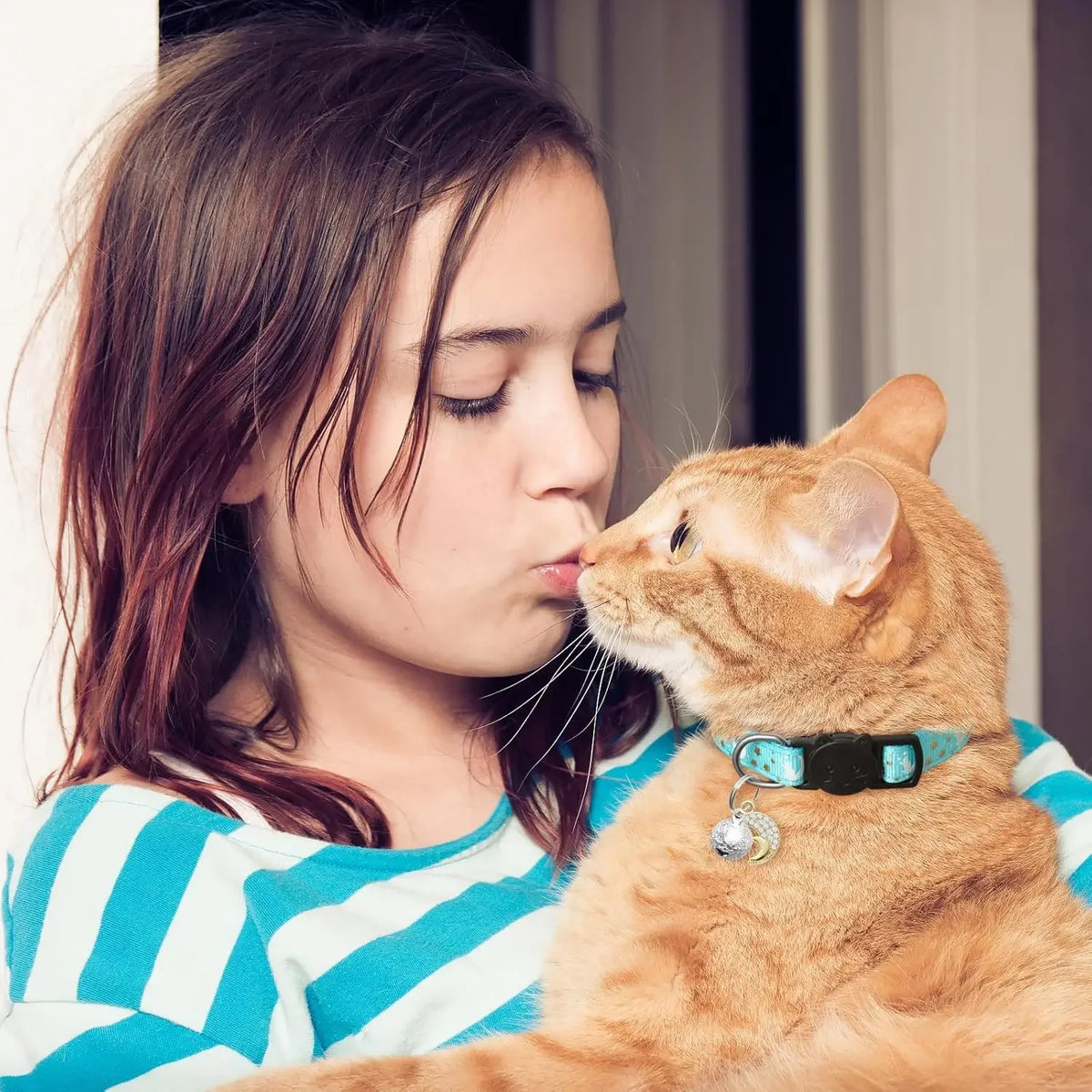 A young girl is gently kissing an orange tabby cat wearing a glow-in-the-dark breakaway collar with a moon pendant and bell.