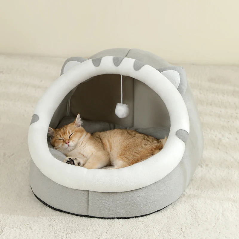 Un gato atigrado naranja y esponjoso está relajándose dentro de una cama de guarida redonda y gris con una abertura blanca.