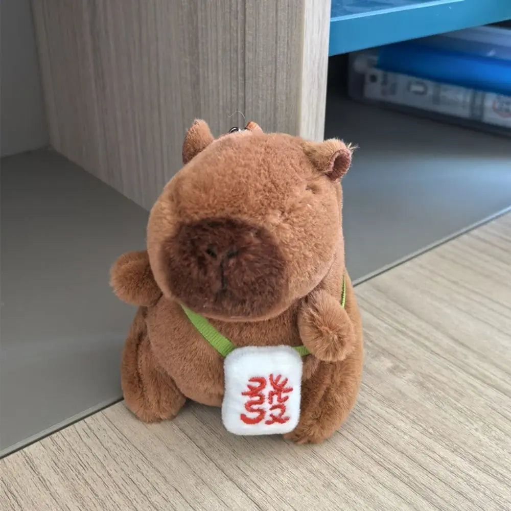 Braune Capybara-Plüschpuppe mit grünem Riemchen und weißem Etikett auf der Brust