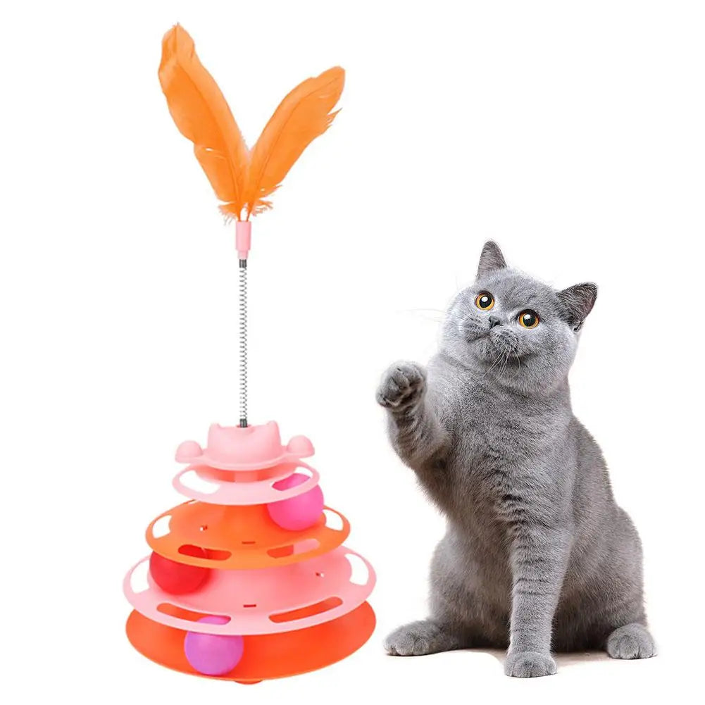A gray cat is sitting next to an orange and pink interactive rotating feather toy tower.