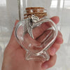 A clear glass heart-shaped jar with a cork stopper and a small silver charm featuring a winged silhouette is held in a hand.