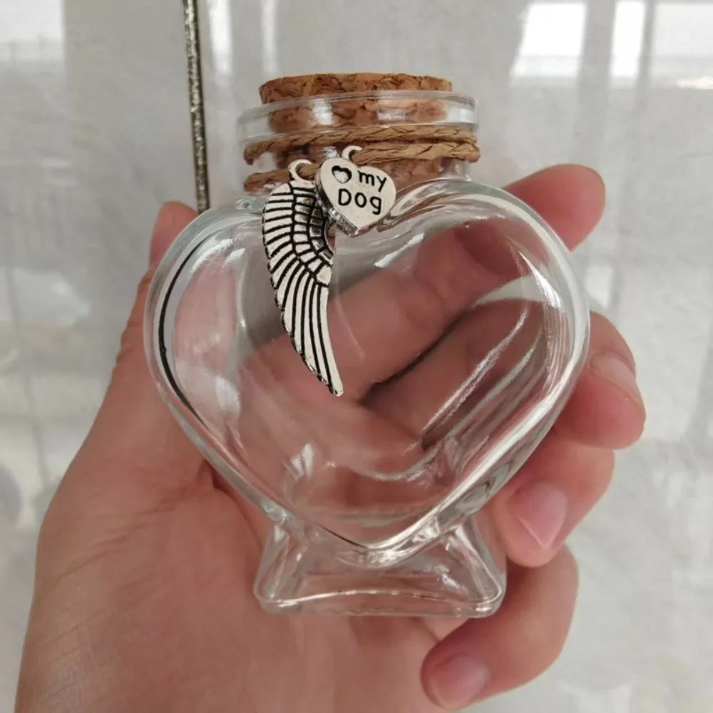 A clear glass heart-shaped jar with a cork stopper and a small silver charm featuring a winged silhouette is held in a hand.