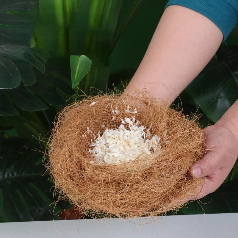 A hand holds a nest made of natural coconut fiber bird bedding against a dark green leaf background.