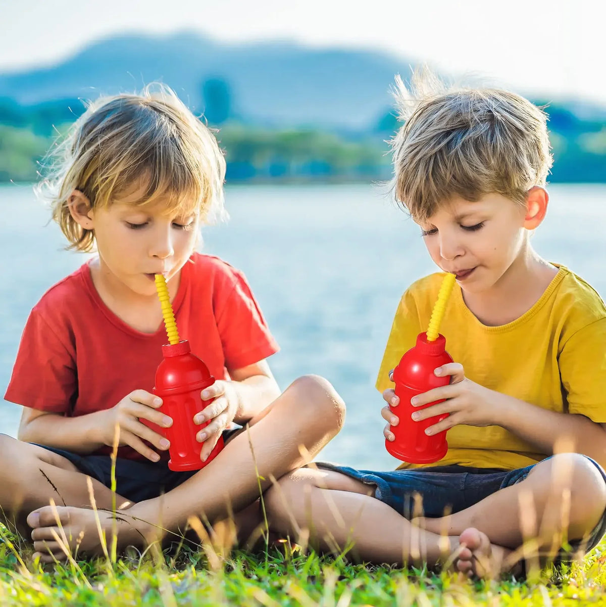 Two young boys are sitting in grass by water, each holding a red fire hydrant-shaped straw cup.