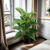 A large artificial monstera plant with green patterned leaves sits in a pot on a wooden floor next to a window.