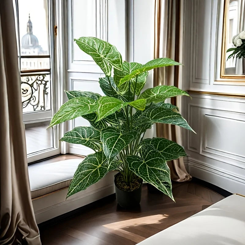 A large artificial monstera plant with green patterned leaves sits in a pot on a wooden floor next to a window.