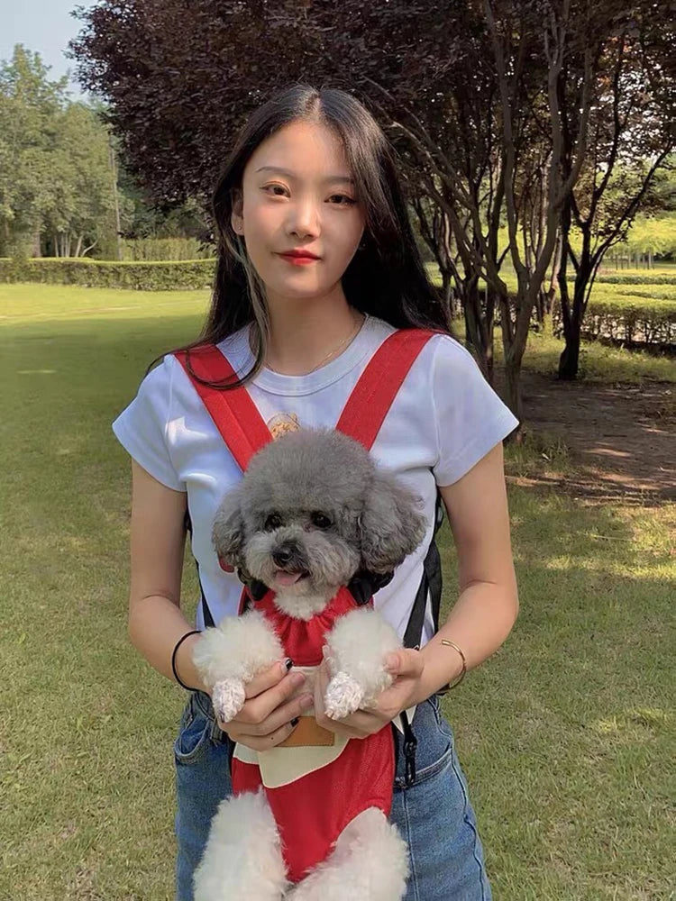 A young woman holds a small, fluffy gray dog in a red pet carrier backpack outdoors.