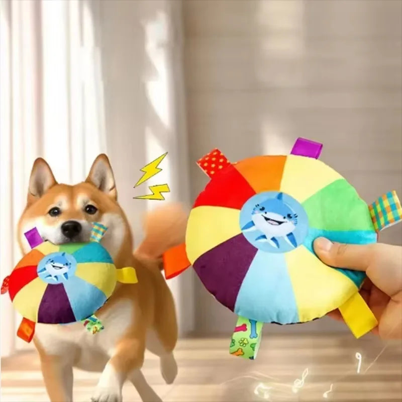 A hand holds a colorful plush duck dog toy with a squeaker, next to an orange Shiba Inu puppy.