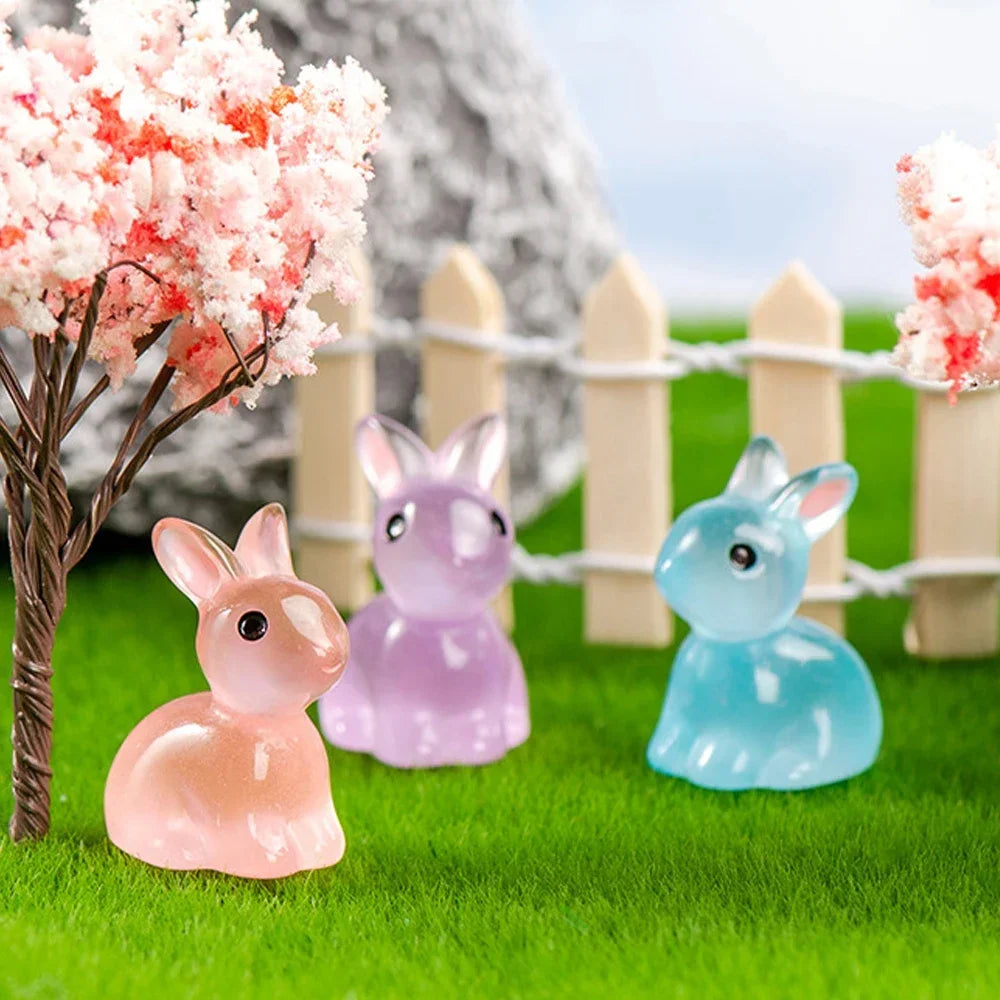 A set of glow-in-the-dark resin bunny figurines in pink and blue colors standing in green grass behind a white picket fence.