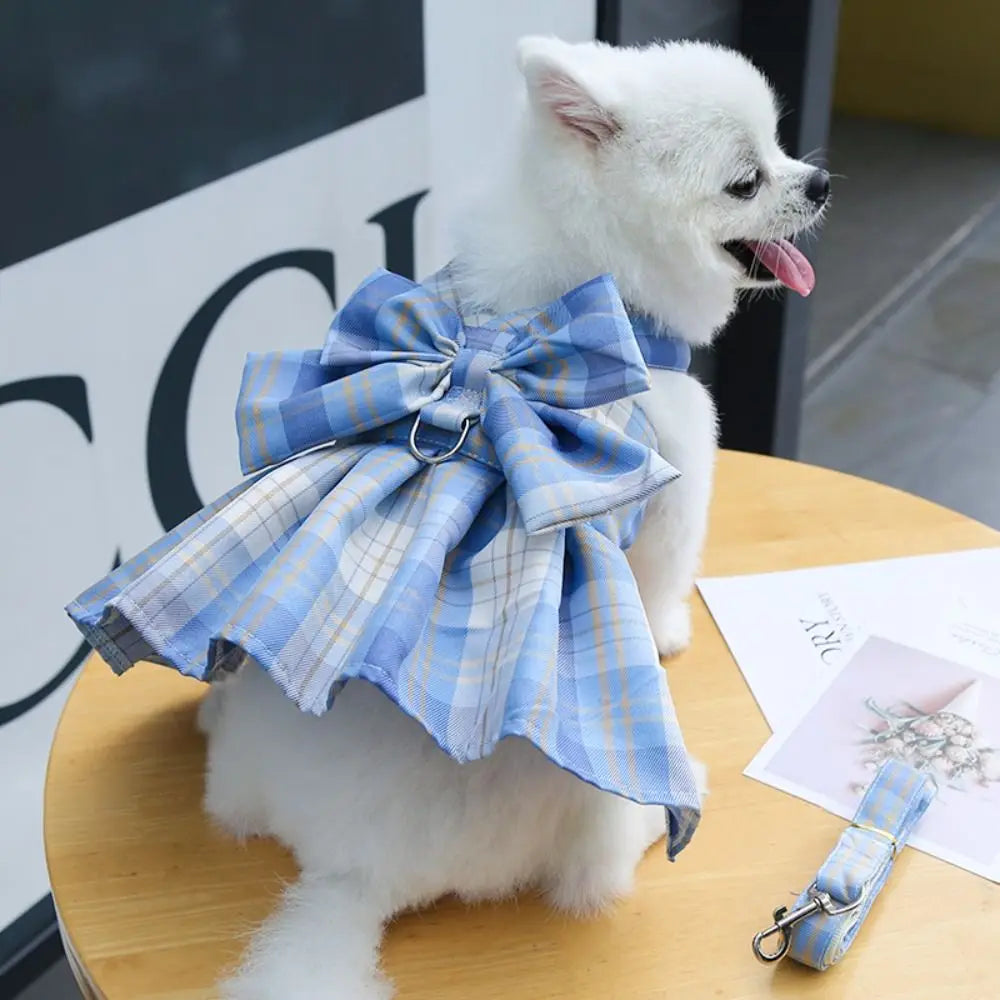 A white dog wearing a blue plaid harness dress with a bow, sitting on a wooden surface.