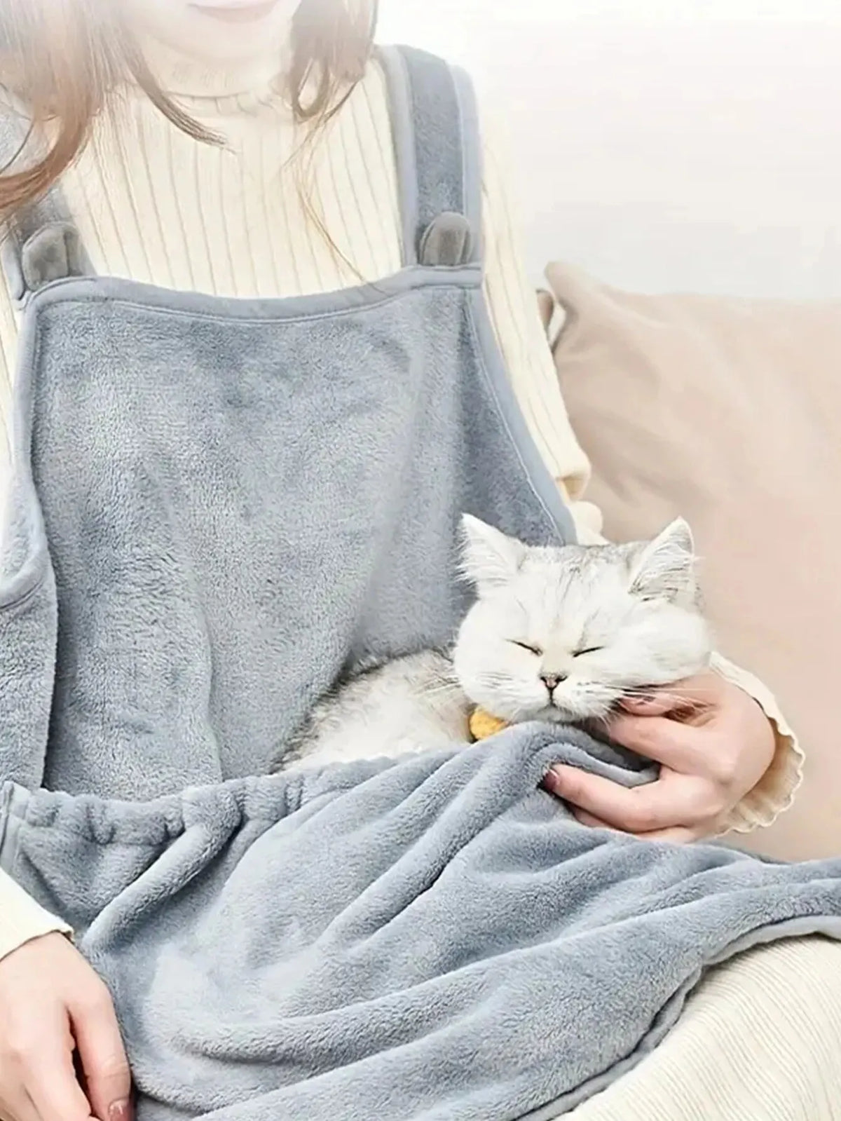 Une personne portant un tablier de transport pour animaux gris et duveteux tient un chat blanc et une couverture assortie gri