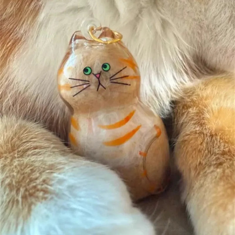 A small, sculpted cat charm keychain with orange stripes and green eyes resting on soft fur.
