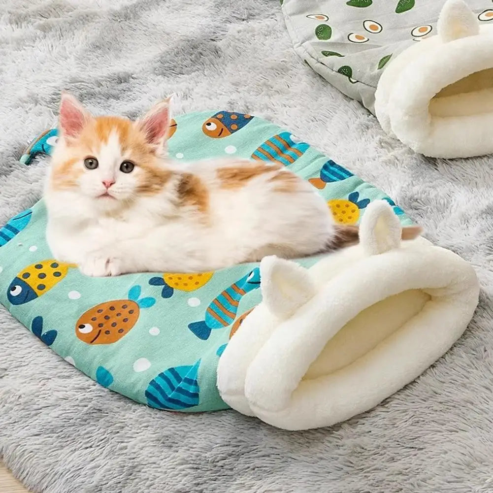 A fluffy orange and white kitten is relaxing inside a cozy plush sleeping bag with a tunnel hideaway on a gray surface.