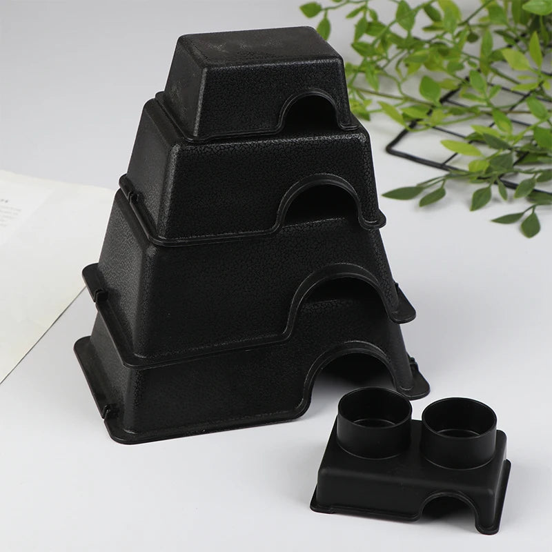 A stack of black reptile hide box enclosures on a white surface with green foliage in the background.
