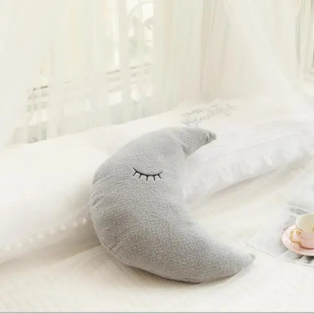 A gray crescent moon shaped plush pillow resting on white bedding near a sheer curtain.