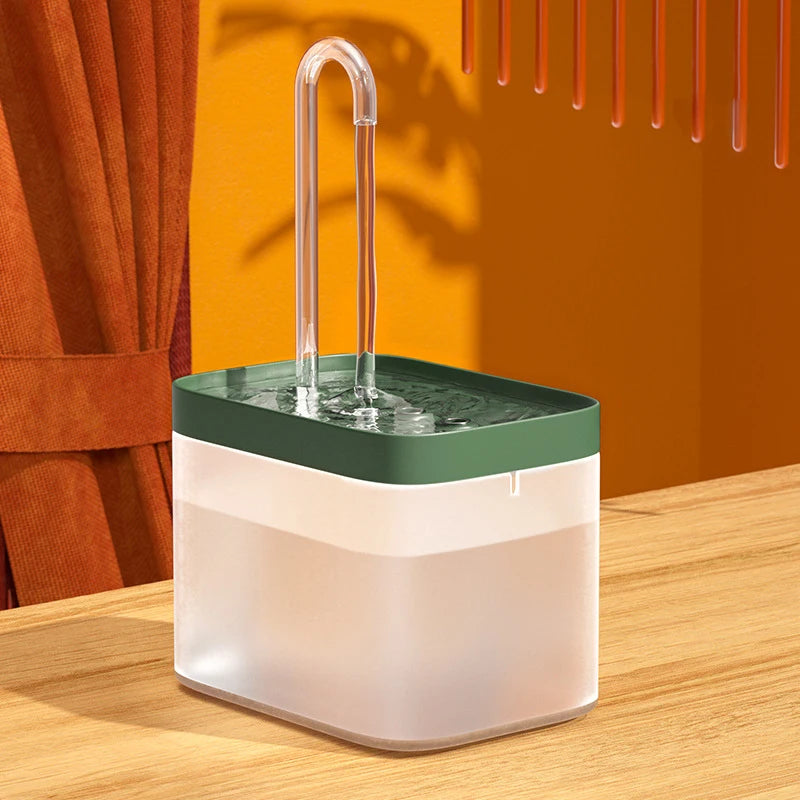A clear pet water fountain with a green base and a stainless steel drinking spout sits on a wooden surface.