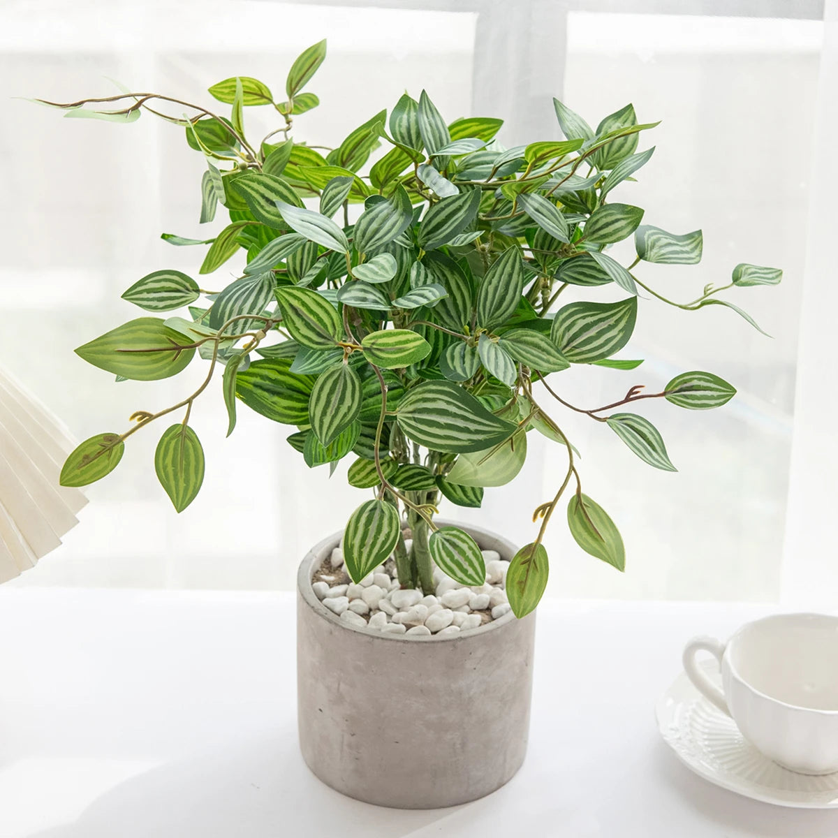 An artificial bonsai tree with green leaves potted in a gray ceramic container on a white surface.