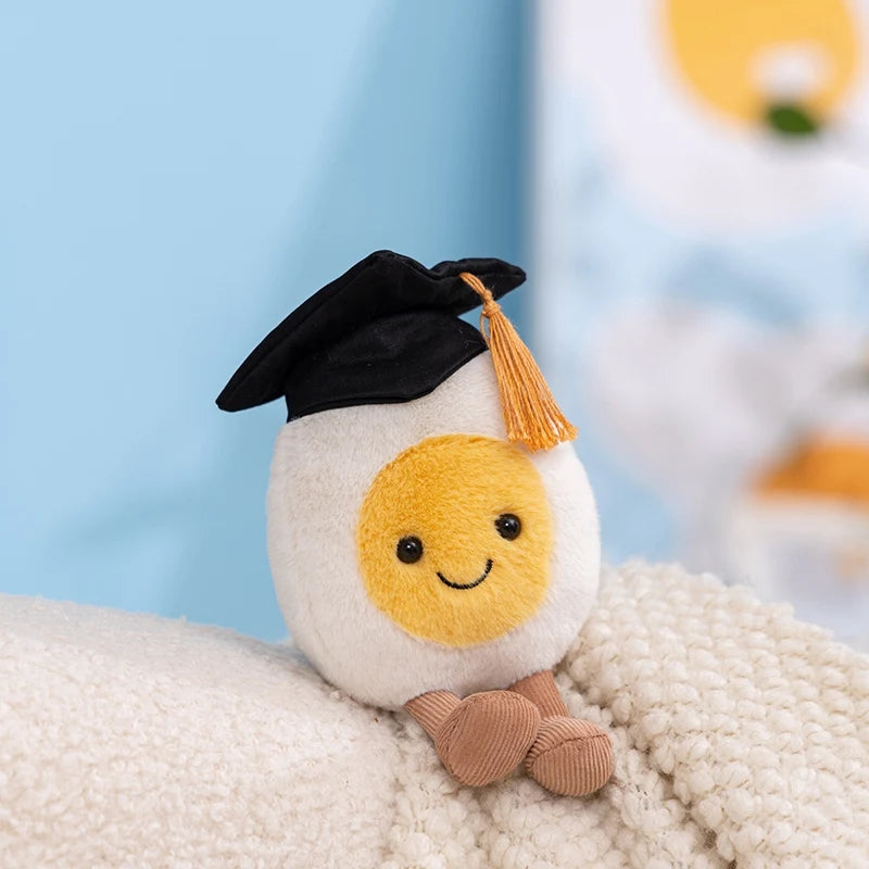 A cute plush toy resembling an egg with a black graduation cap and a yellow face, resting on a white textured surface.