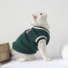A white cat wearing a dark green sweater with a black trim and a small patch on its chest.