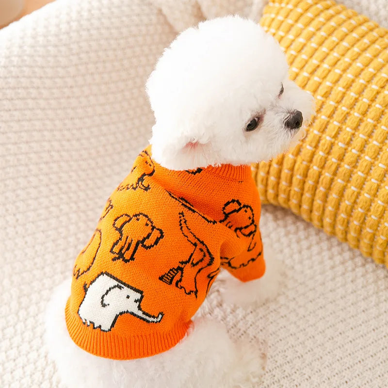 A white dog wearing an orange knit sweater with elephant patterns, resting on a cream-colored blanket.