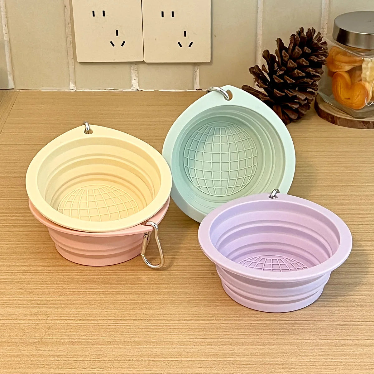 Three collapsible silicone pet travel bowls in pale yellow, mint green, and lavender are displayed on a wooden surface.