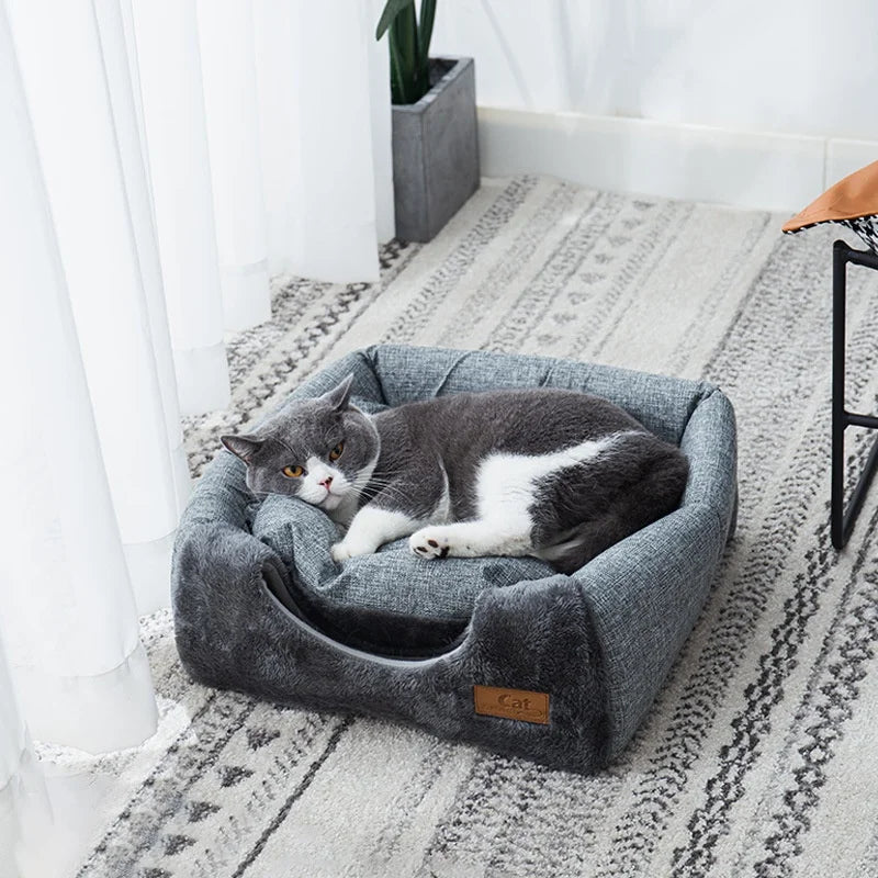 Un lit-nid de chat en velours gris est placé sur un tapis clair près d'une fenêtre.