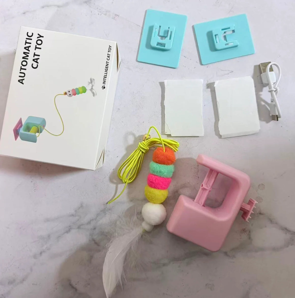 An automatic electric hanging cat toy with a cord and a pink plastic component is shown on a white marble surface.